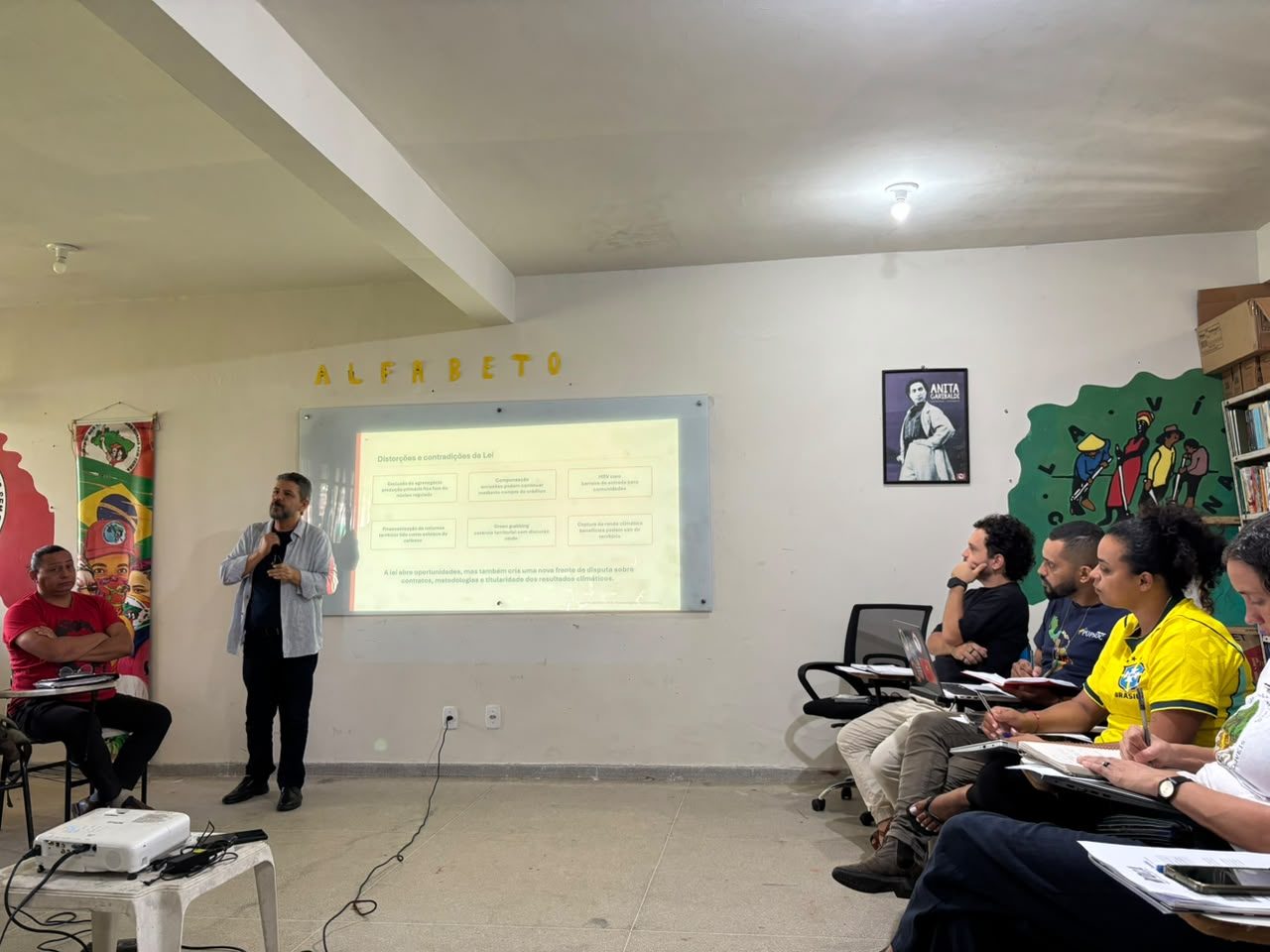 Pessoa apresentando ao microfone para uma plateia, com projeção ao fundo